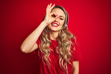 Young beautiful woman wearing headphones over red isolated background doing ok gesture with hand smiling, eye looking through fingers with happy face.