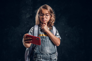 Little girl in glasses is scrolling mobile phone at dark photo studio.