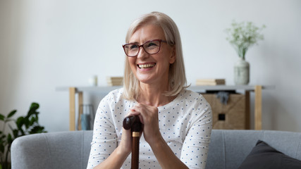 Smiling mature woman holding wooden cane, sitting on couch