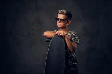 Young schoolboy in sunglasses is posing at dark photo studio with his skateboard.