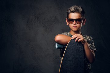 Young schoolboy in sunglasses is posing at dark photo studio with his skateboard.