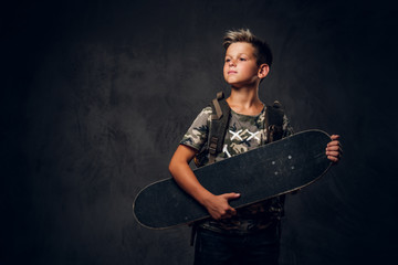 Small attractive boy is posing at dark photo studio with his skateboard on the dark background.