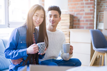 Young asian couple drinking a cup of coffee relaxing on the sofa at new home, smiling happy around cardboard boxes from moving