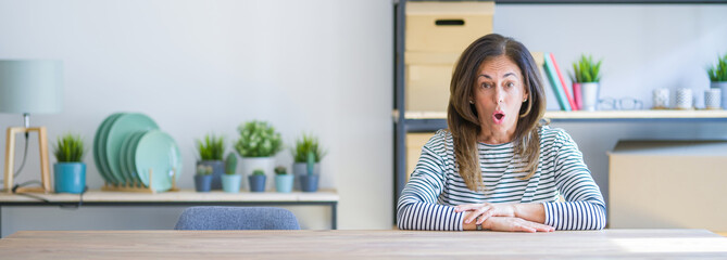 Wide angle photo of middle age senior woman sitting at the table at home afraid and shocked with surprise expression, fear and excited face.