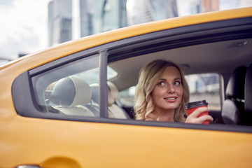 Picture of happy blonde with glass of coffee in her hands sitting in back seat of yellow taxi.