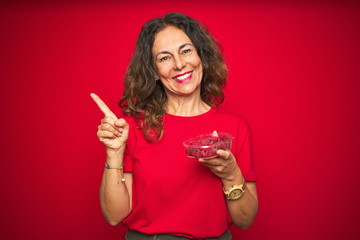 Naklejka premium Middle age senior woman eating raspberries over red isolated background very happy pointing with hand and finger to the side
