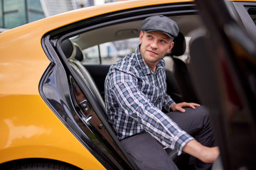 Image of man in cap and plaid shirt looking to side sitting in back seat in yellow taxi