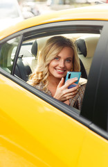 Photo of young blonde woman with phone in her hand sitting in back seat of yellow taxi.