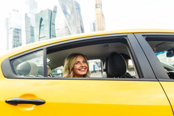 Photo of young woman sitting in back seat of yellow taxi in summer on modern city background.
