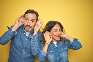 Beautiful middle age couple together standing over isolated yellow background Trying to hear both...