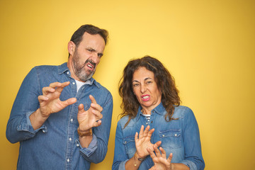 Beautiful middle age couple together standing over isolated yellow background disgusted expression, displeased and fearful doing disgust face because aversion reaction. With hands raised. 