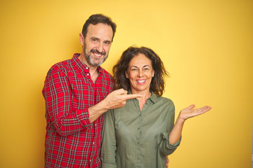 Fototapeta premium Beautiful middle age couple over isolated yellow background amazed and smiling to the camera while presenting with hand and pointing with finger.