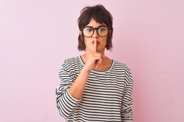 Young beautiful woman wearing striped t-shirt and glasses over isolated pink background asking to be quiet with finger on lips. Silence and secret concept.