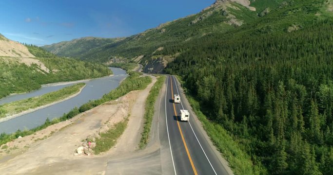 Alaska. Aerial 4K. Camera follows RVs driving along Alaskan mountain road. Calm mountain river is on the left and dense vegetation is on the right of the way. The bends of the road are seen ahead.