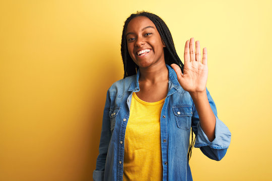 Young african american woman wearing denim shirt standing over isolated yellow background Waiving saying hello happy and smiling, friendly welcome gesture