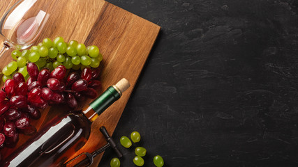 White wine bottle, bunch of grapes and wineglass on wooden board and black background