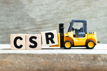 Toy forklift hold letter block r to complete word CSR (Abbreviation of corporate social responsibility) on wood background