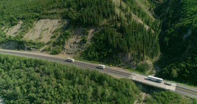 Alaska. 4K. Aerial side view on the caravan of three white Rvs moving one after another along the way among the mountains. The slopes are covered with trees. The scenery is lit by the sun.