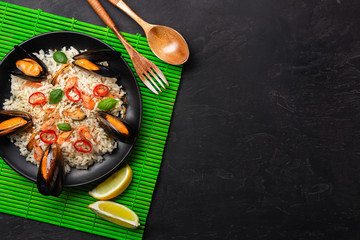 Fried rice with seafood mussels, shrimps, basil in a black plate with wooden spoon and fork on green bamboo mat and stone table