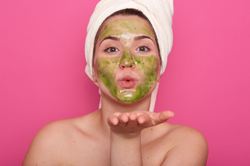 Beauty portrait of relaxed Caucasian woman in spa salon with green facial mask, wearing white towel isolated over rose background, charming female showing kiss gesture, looking directly at camera.