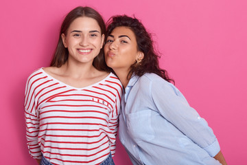 Close up portrait of two young hipster girls, best friends, wearing casual shirts, having great time togather, female with wavy hair making kiss gesture, women posing isolated over pink background.