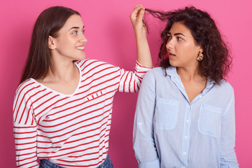 Close up portrait of two young women, girl wearing striped shirt touches friends hair with fingers, attractive lady looks at beautiful lady with shocked facial expression, isolated on rose background.