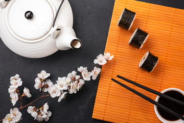 Set of sushi and maki rolls with branch of white flowers and teapot on stone table