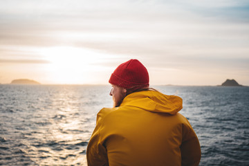 Alone man traveling on ship and looking at sunset sea and foggy mountain on skyline. Hipster traveler wearing yellow raincoat and red hat enjoying beautiful ocean after storm - obrazy, fototapety, plakaty