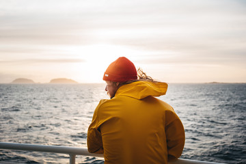 Hipster traveler wearing red hat and yellow raincoat looking away at cloudy mountain and sunset sea. Alone man traveling  at scandinavian authentic ocean landscape by ship