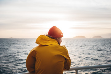 Young traveler wearing red hat and yellow raincoat floating on ship looking at sunset sea after storm and foggy mountains on skyline. Lifestyle travel, scandinavian authentic landscape.