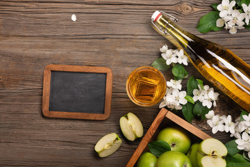 Ripe green apples in wooden box with branch of white flowers, glass, bottle of fresh juice and chalk board on a wooden table