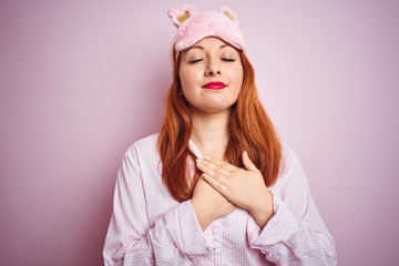 Young beautiful redhead woman wearing pajama over pink isolated background smiling with hands on chest with closed eyes and grateful gesture on face. Health concept.
