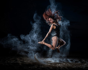 Fototapeta na wymiar Ballet dancer in black bodysuit jumps high and dance with flour on the beach in the evening. Women's street ballet. Flour, talc, dust. Young red-haired woman jumping in a cloud of blue powder.
