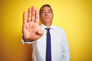 Handsome middle age businessman standing over isolated yellow background doing stop sing with palm...
