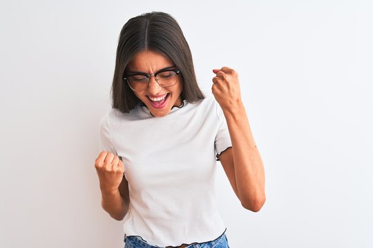 Young beautiful woman wearing casual t-shirt and glasses over isolated white background very happy and excited doing winner gesture with arms raised, smiling and screaming for success. 