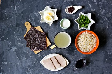 Ingredients for making ramen soup on a dark concrete background: prepared ramen noodles, pork, miso paste, soy sauce, broth, chopped green onions, boiled egg, sugar, nori seaweed. Japanese food.