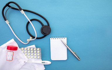 a stethoscope next to a medical gown lie on a blue background next to a thermometer with a notepad and pen