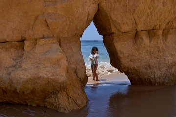 Niña rizosa morena caminando por la playa con los pies en el agua entre las formaciones rocosas