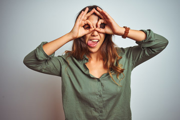 Young beautiful woman wearing green shirt standing over grey isolated background doing ok gesture like binoculars sticking tongue out, eyes looking through fingers. Crazy expression.