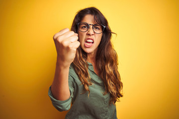 Young beautiful woman wearing green shirt and glasses over yelllow isolated background angry and mad raising fist frustrated and furious while shouting with anger. Rage and aggressive concept.