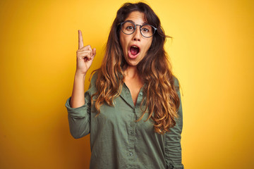 Young beautiful woman wearing green shirt and glasses over yelllow isolated background pointing finger up with successful idea. Exited and happy. Number one.