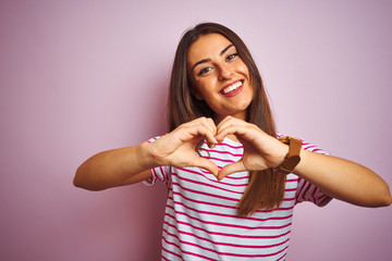 Young beautiful woman wearing striped t-shirt standing over isolated pink background smiling in love showing heart symbol and shape with hands. Romantic concept.