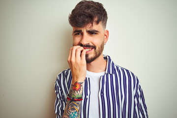 Young man with tattoo wearing striped shirt standing over isolated white background looking stressed and nervous with hands on mouth biting nails. Anxiety problem.