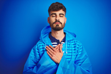 Young handsome man wearing rain coat standing over isolated blue background smiling with hands on chest with closed eyes and grateful gesture on face. Health concept.