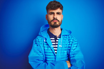 Young handsome man wearing rain coat standing over isolated blue background skeptic and nervous, disapproving expression on face with crossed arms. Negative person.