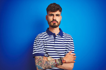 Young man with tattoo wearing striped polo standing over isolated blue background skeptic and nervous, disapproving expression on face with crossed arms. Negative person.