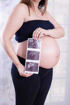 Pregnant woman hold photo or picture on the belly or ultrasound scan on stomach of future daughter or son at home. Girl expecting newborn. Motherhood concept. Baby Shower.