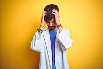 Young doctor man with tattoo standing over isolated yellow background suffering from headache desperate and stressed because pain and migraine. Hands on head.