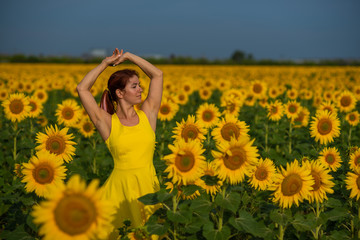 Fototapeta premium A red-haired woman in a yellow dress is standing in a field of sunflowers. Beautiful girl in a skirt sun enjoys a cloudless day in the countryside. Pink locks of hair.