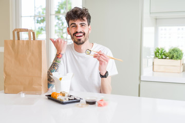 Young man eating asian sushi from home delivery pointing and showing with thumb up to the side with happy face smiling